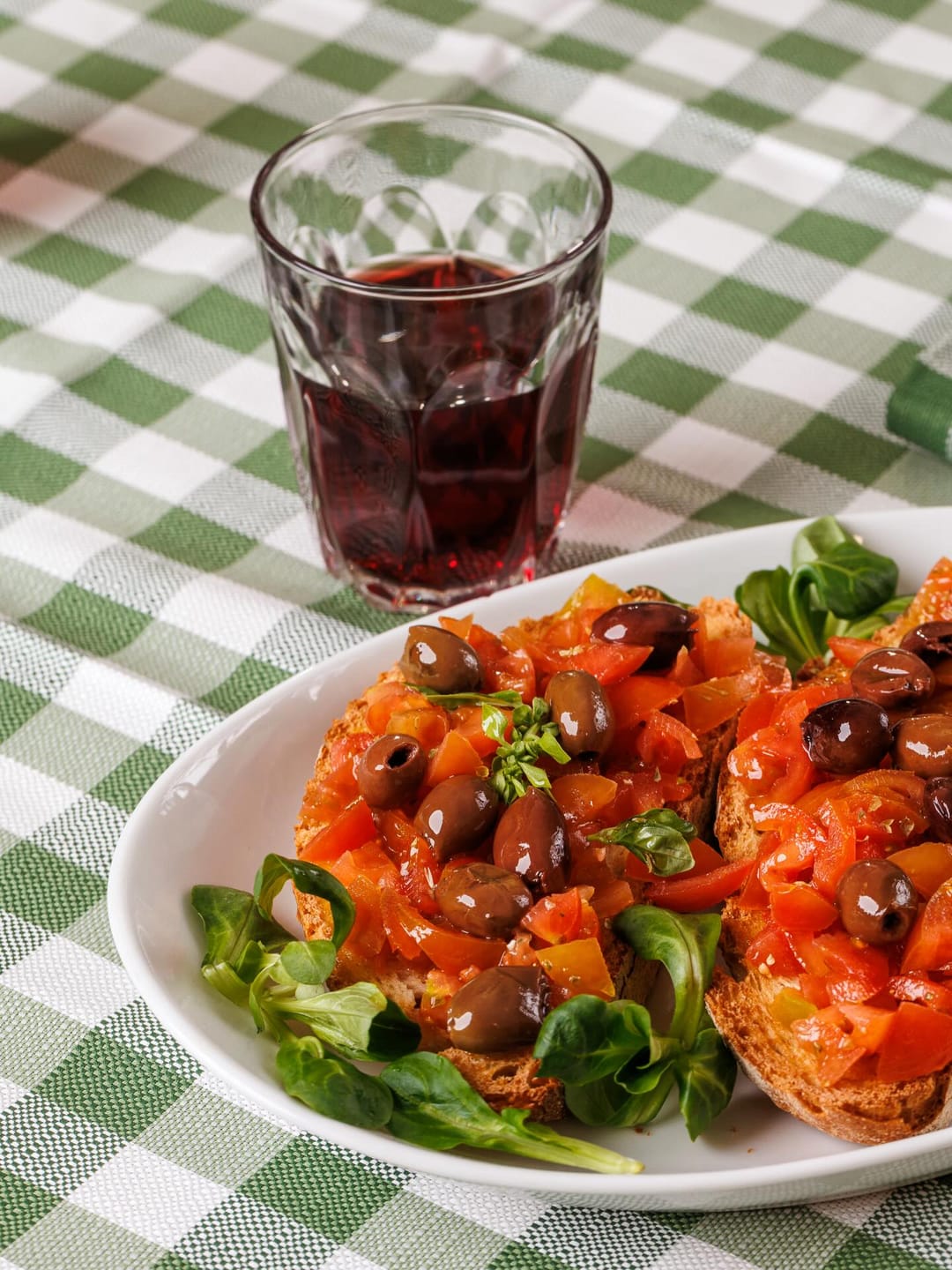 Bruschetta con pomodorini e olive su un piatto bianco, accanto a un bicchiere di vino rosso, su una tovaglia a quadri.