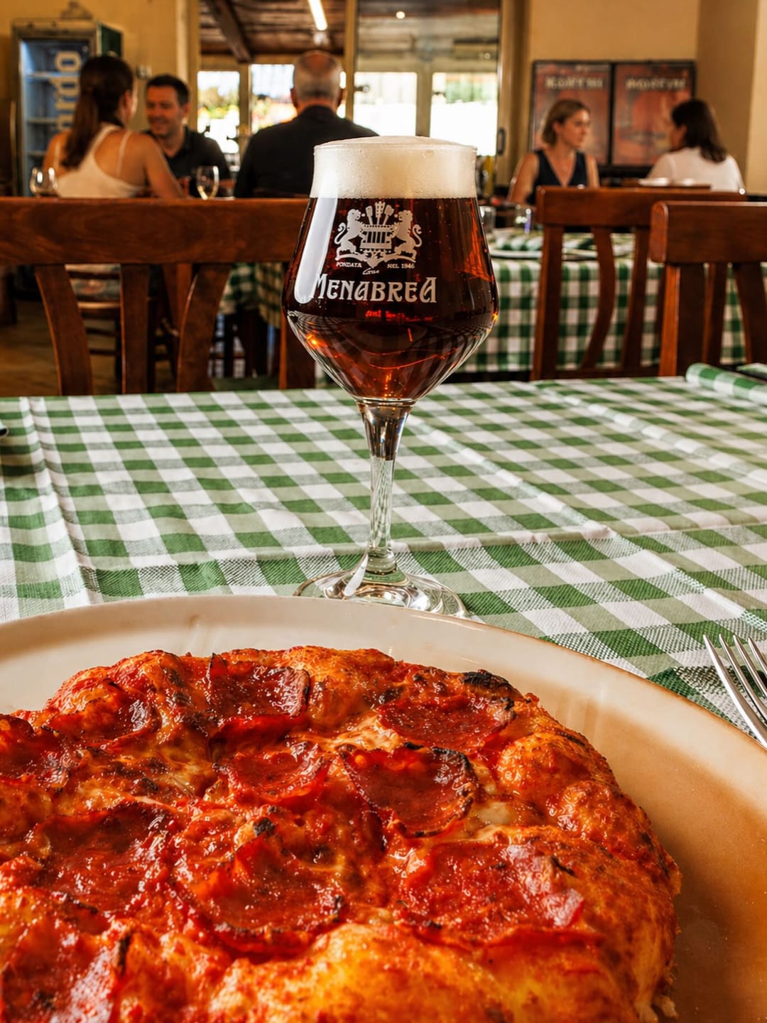 Pizza al padellino con una birra Menabrea su una tovaglia a quadri; sullo sfondo la sala con i clienti.