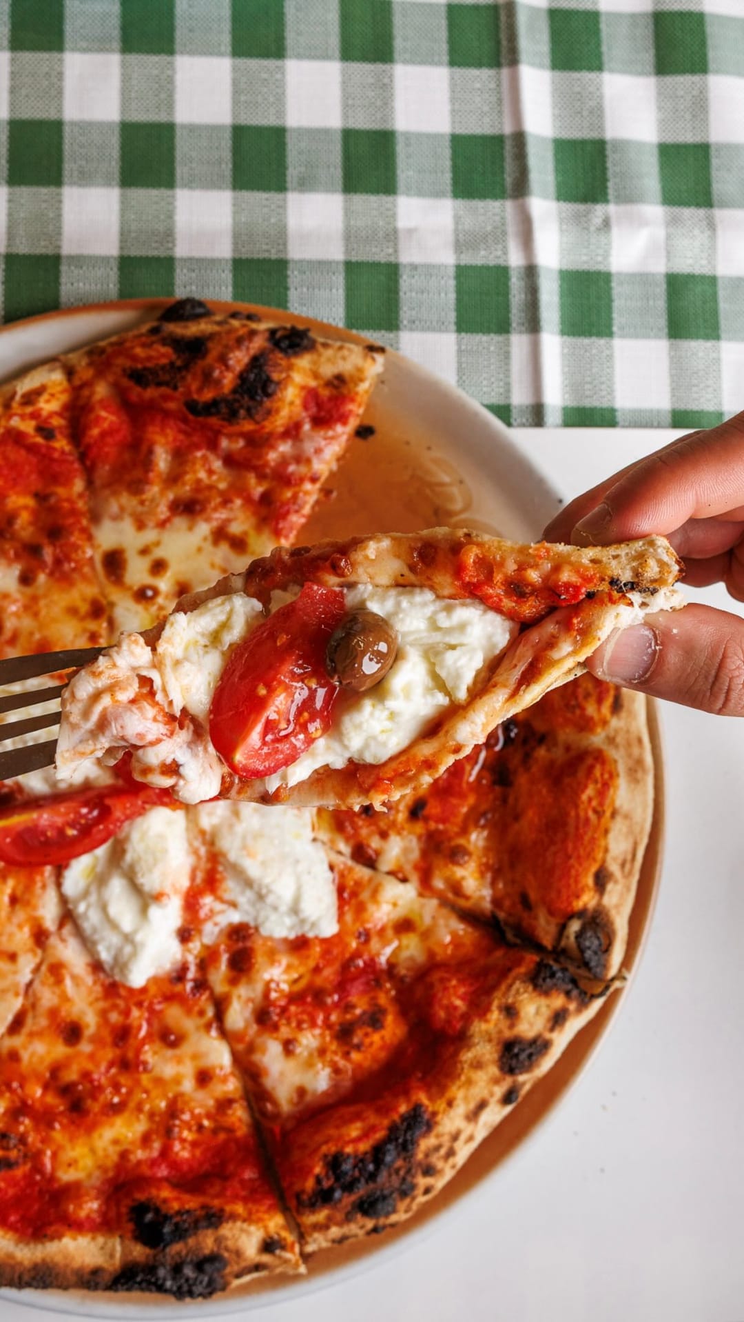 Una fetta di pizza con pomodoro e stracciatella sollevata con la forchetta, su una tovaglia a quadri.