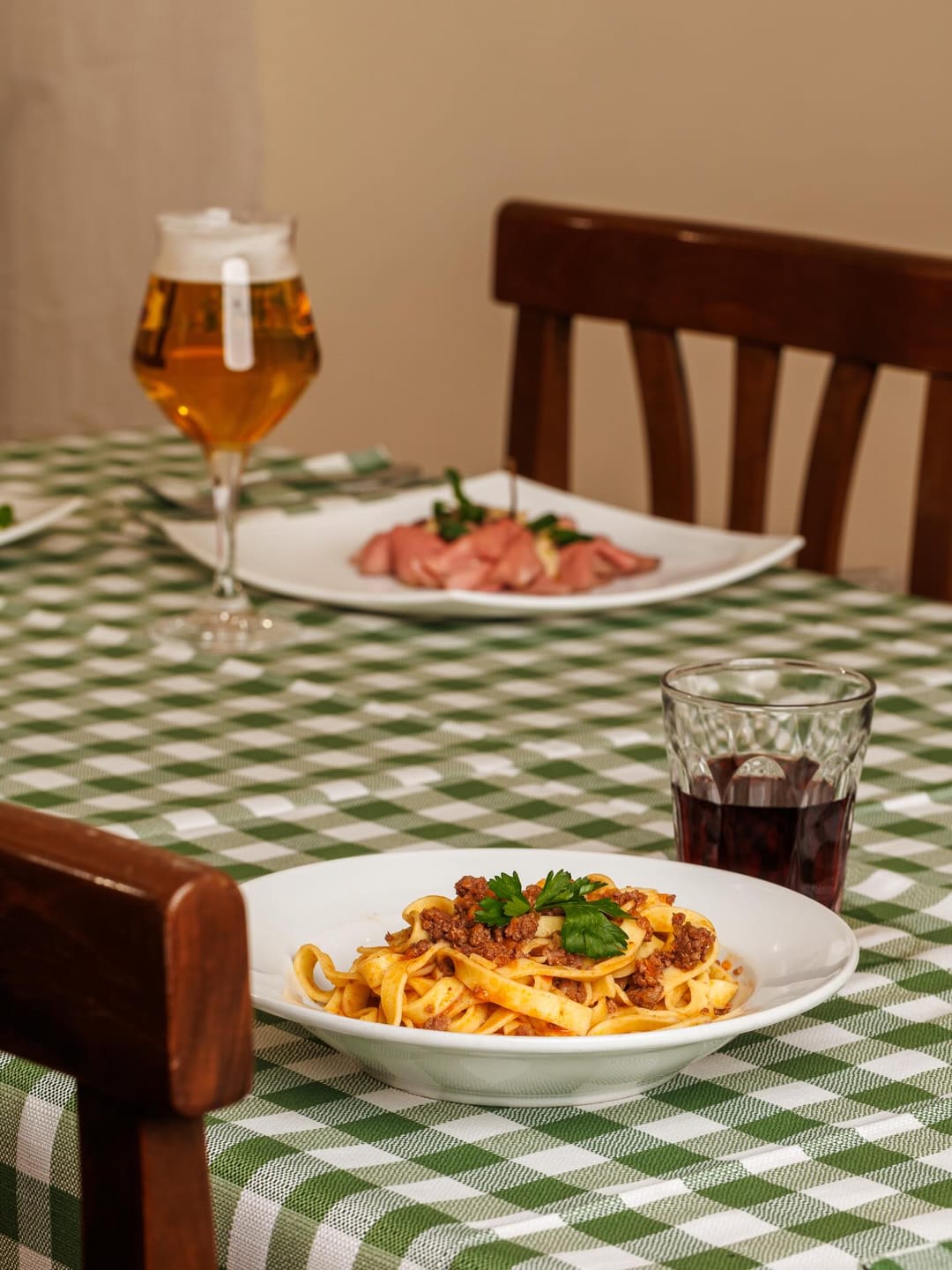 Tagliatelle al ragù su una tavola con tovaglia a quadri, sedie in legno e un calice di birra.