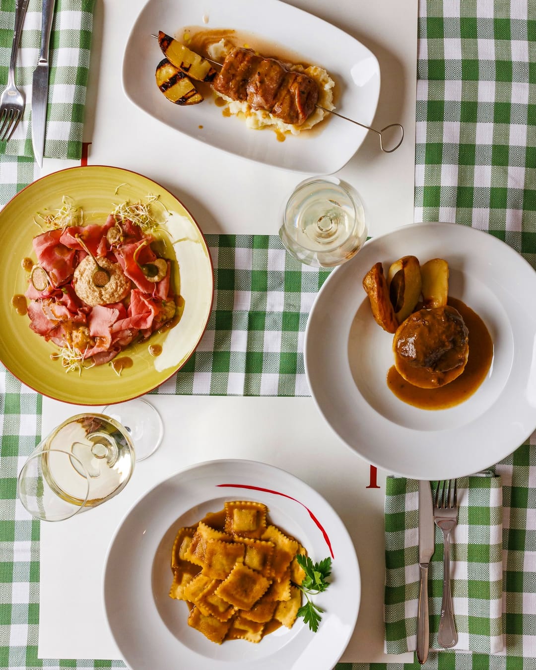 Tavola apparecchiata vista dall’alto, su tovaglia a quadri verdi: vitello tonnato, spiedino con patate, guancia con patate e plin al sugo d’arrosto, con due calici di vino bianco.